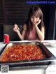 A woman sitting at a table with chopsticks in front of a large pan of food.