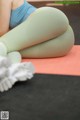 A woman in a blue top and green leggings is sitting on a yoga mat.